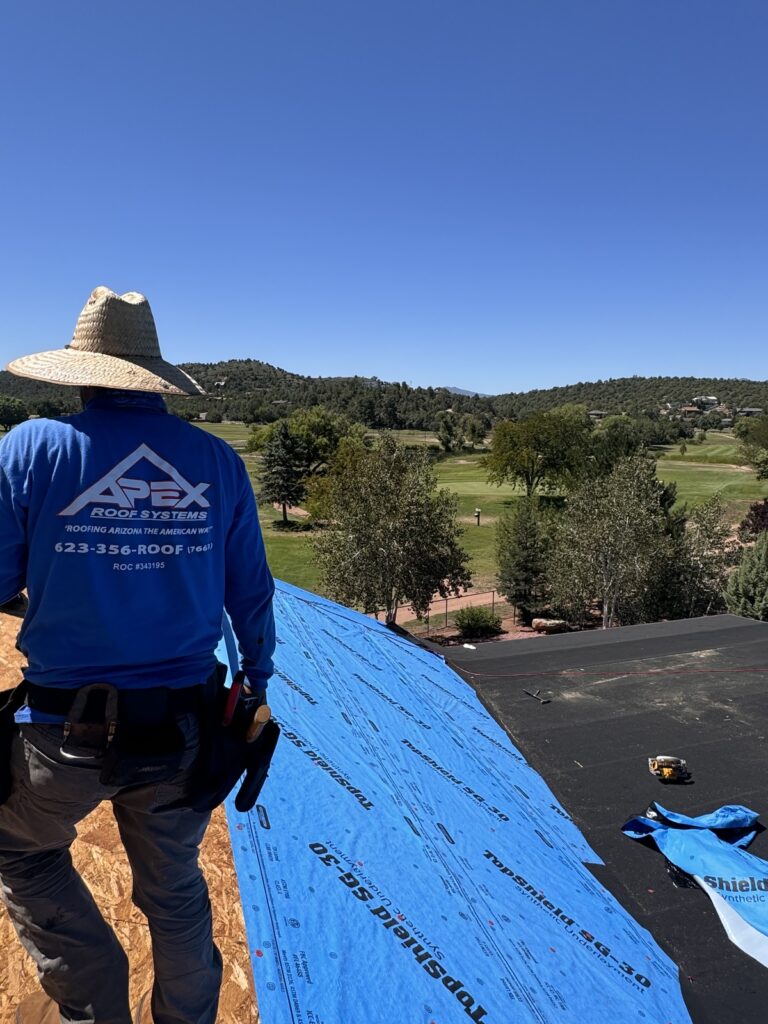 A roofing contractor from Apex Roofing Systems installing a new roof underlayment with a scenic Arizona landscape in the background.